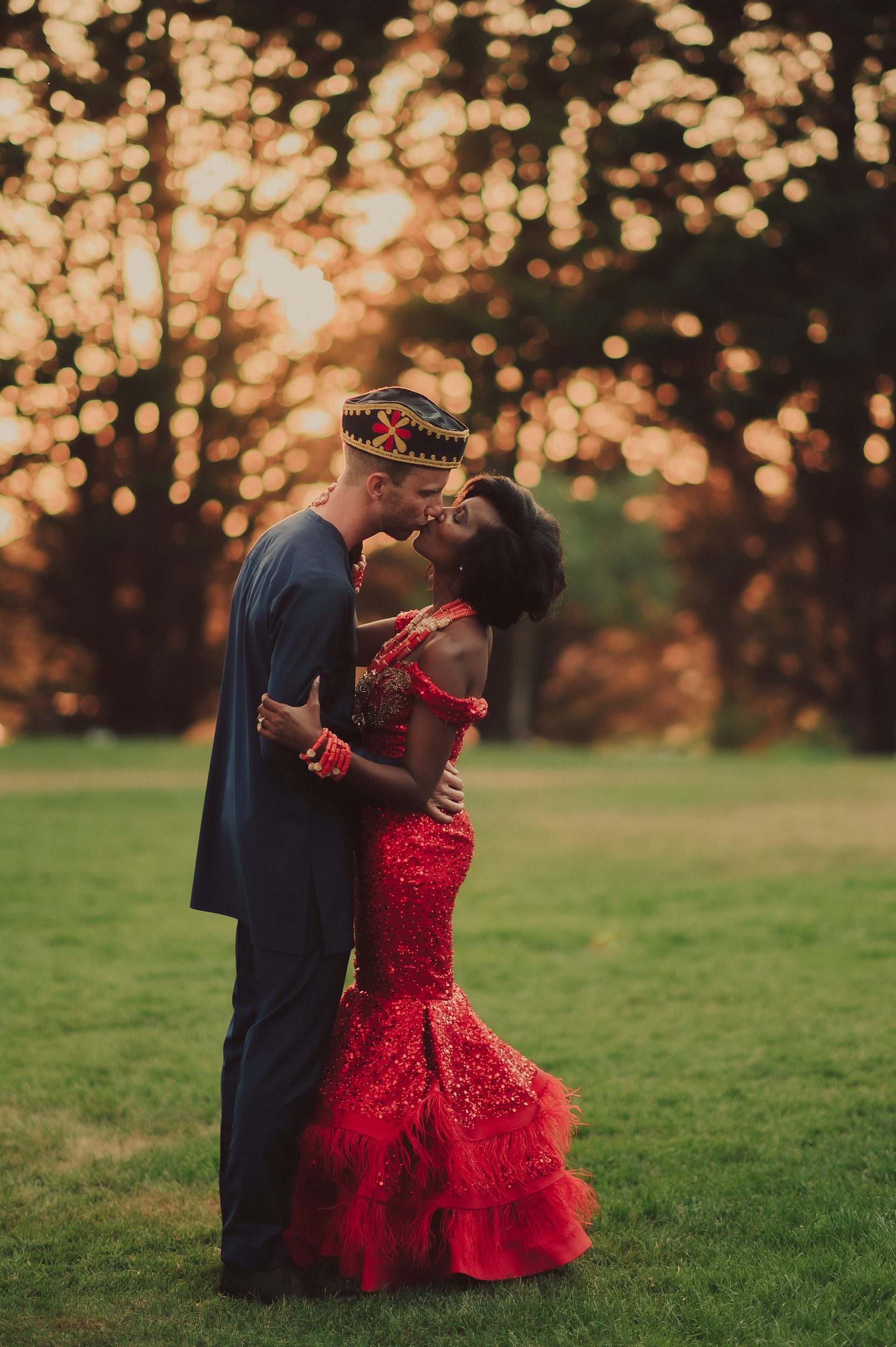 Tracy and Alex share a kiss in red and navy wedding attire as the sun filters through the trees at Heritage Estates.