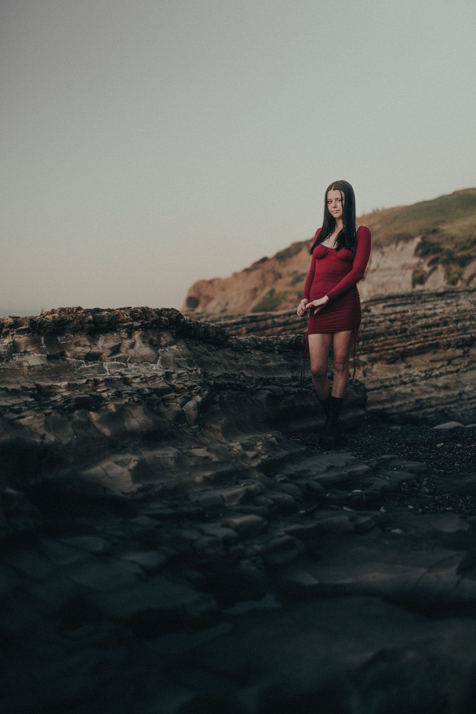 Full-length senior portrait in a red dress on layered coastal rocks