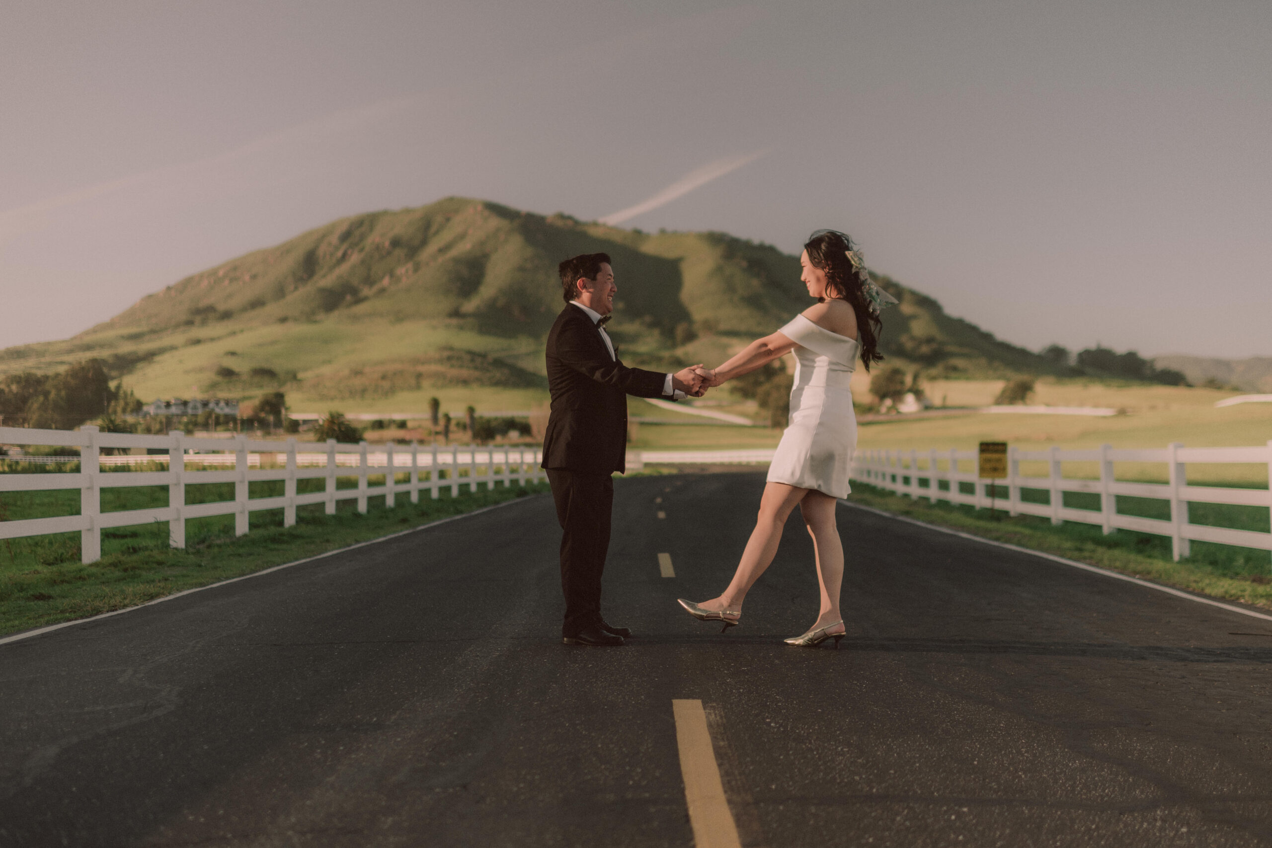 A newlywed couple dancing together on a country road with rolling hills behind them