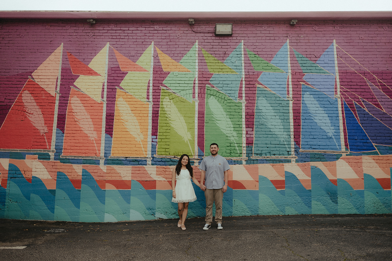 Engaged couple posing together in front of a colorful sailboat mural