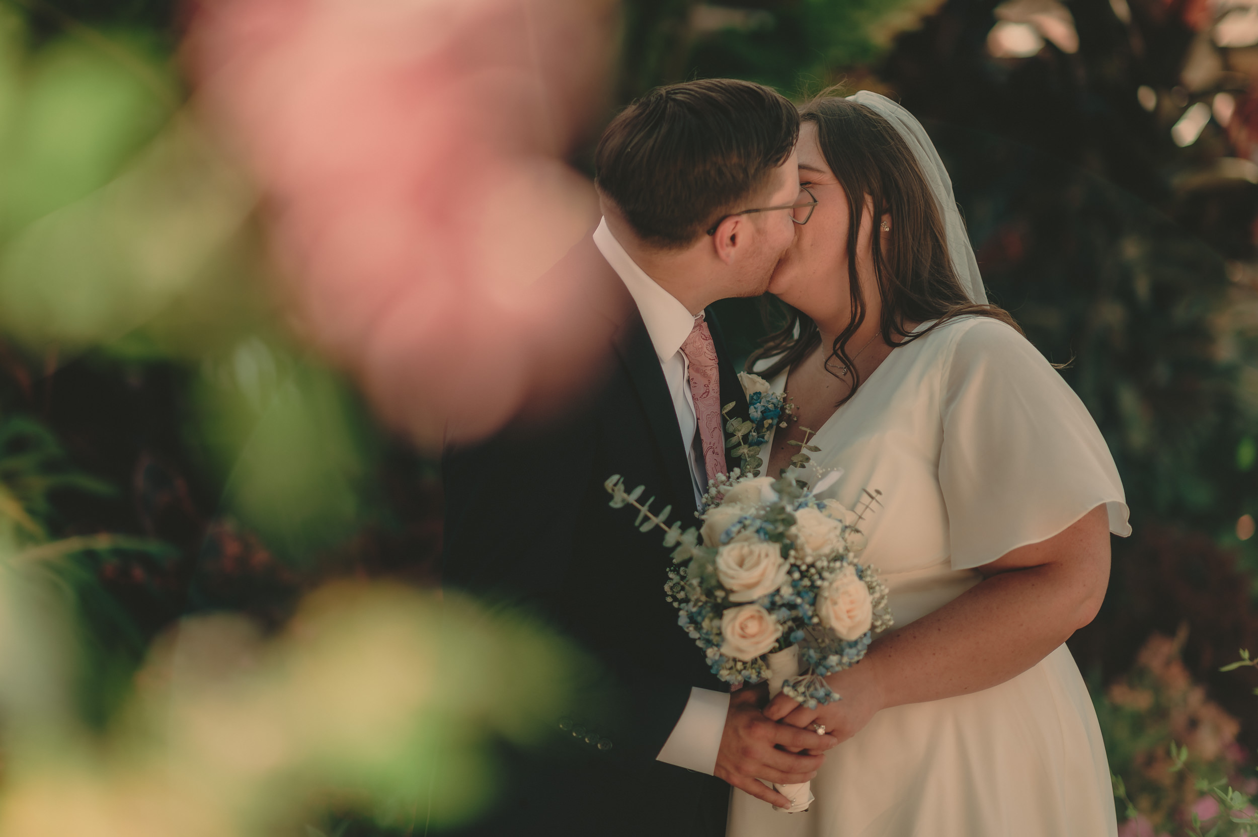 Tiffani and Braden kiss behind pink and green foreground blur.