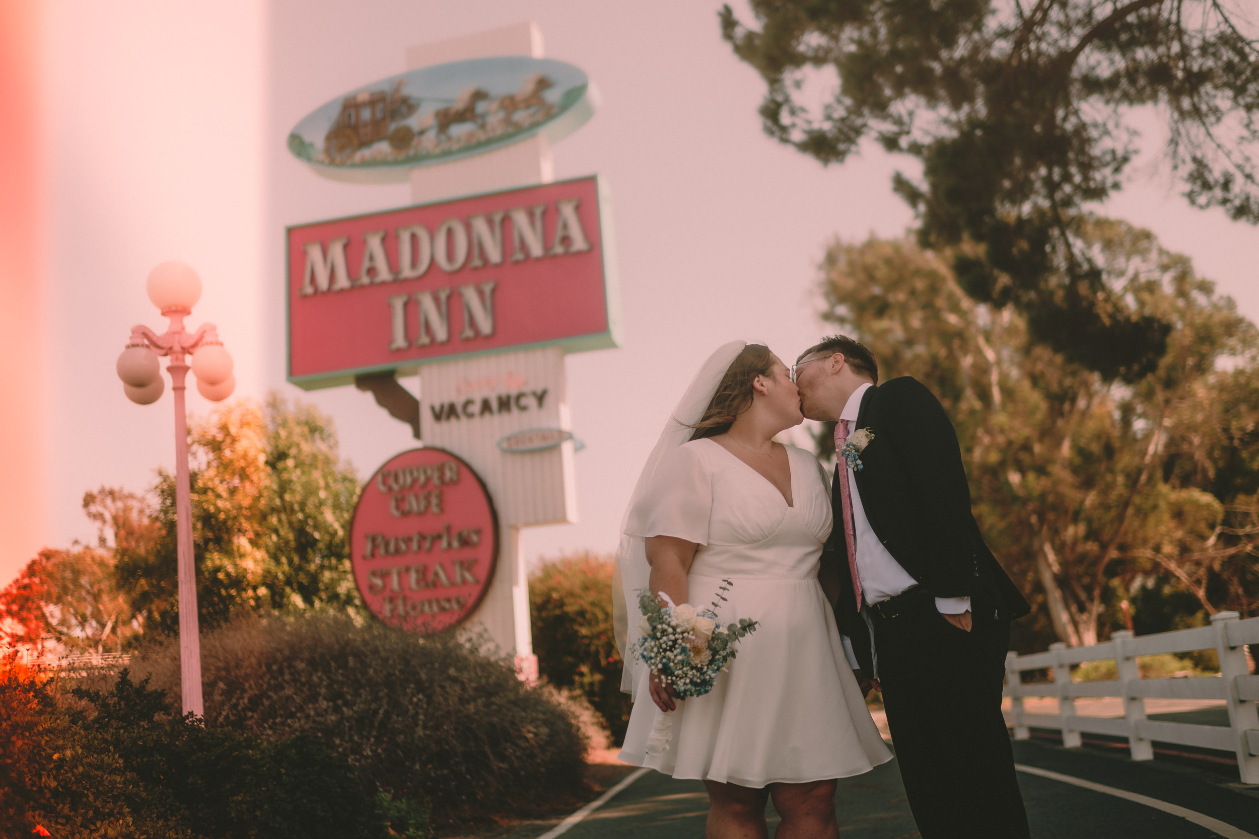 Tiffani and Braden kiss beneath the Madonna Inn sign as a warm light leak crosses the photograph.