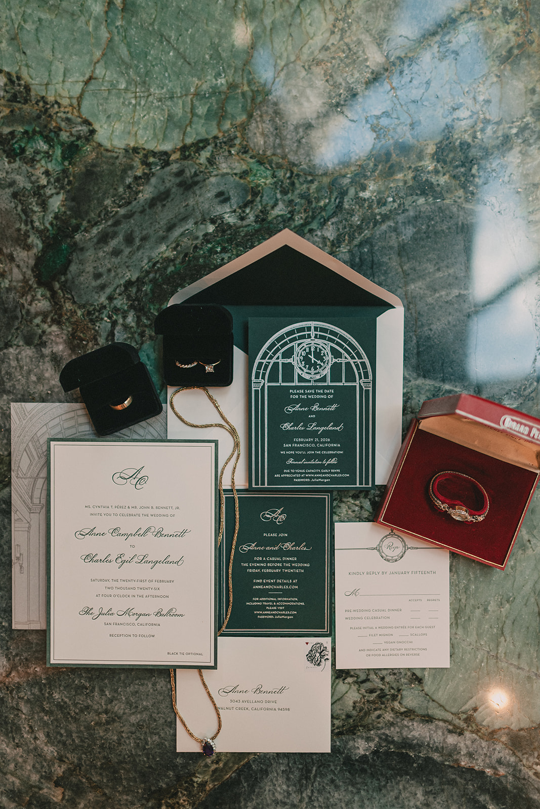 Green and ivory wedding stationery, jewelry and rings arranged on green marble.