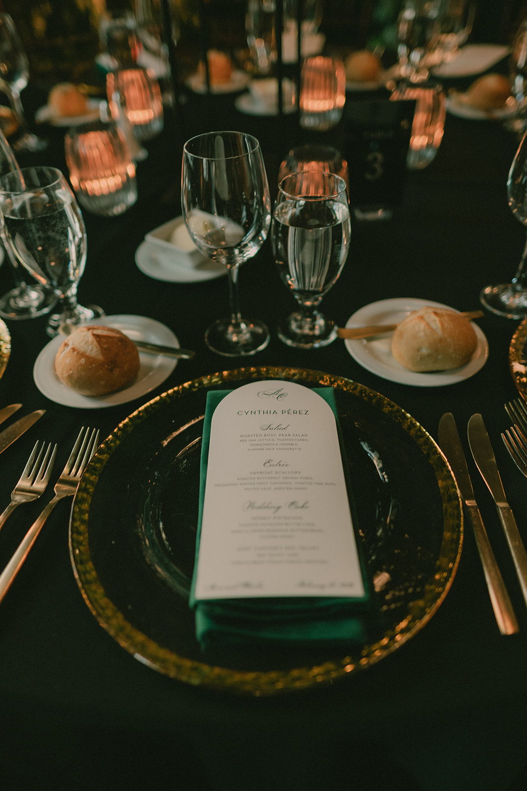 Candlelit place setting with a deep-green napkin, dark glass charger and wedding menu.
