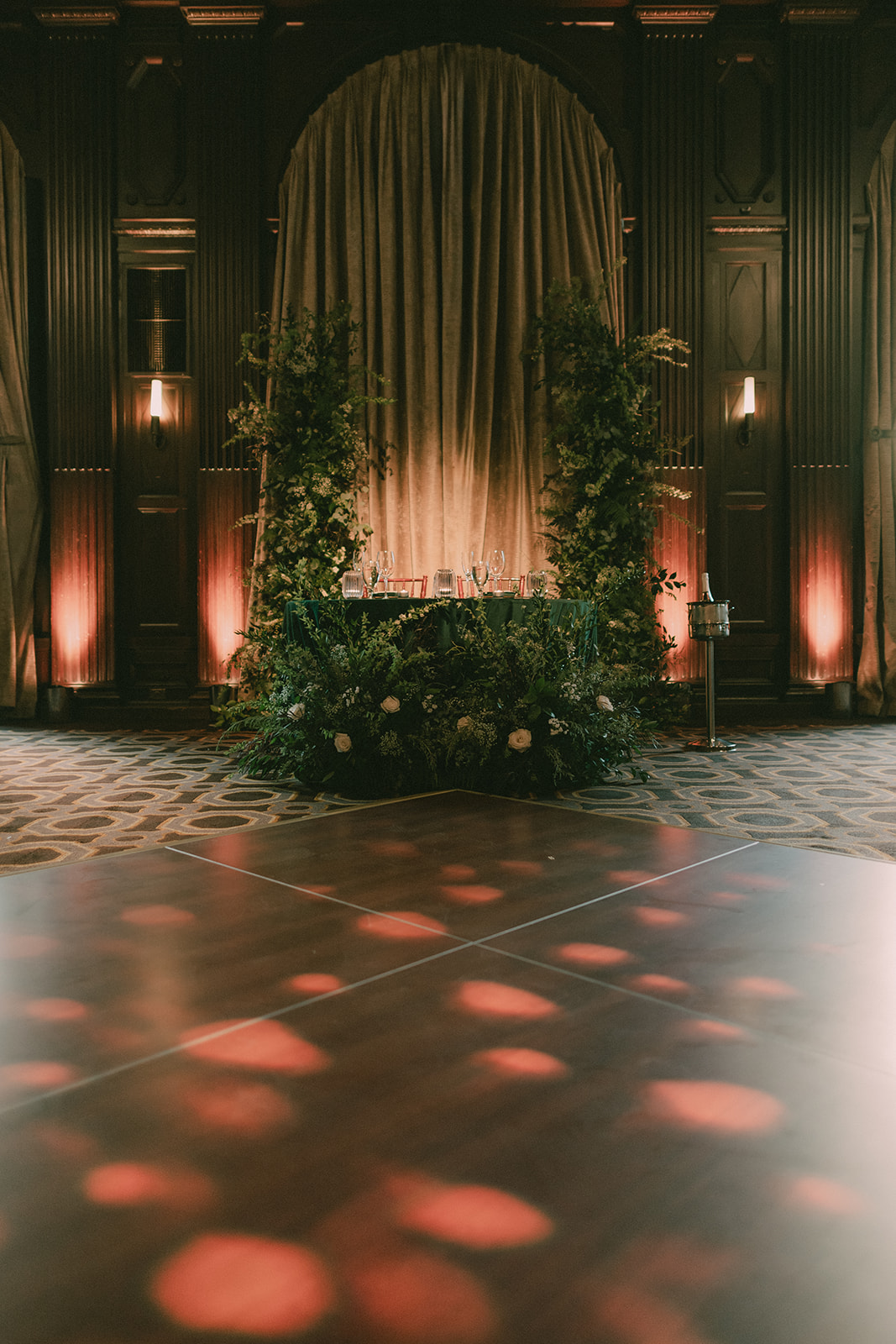 Patterned warm light falls across the empty dance floor and greenery-covered sweetheart table.