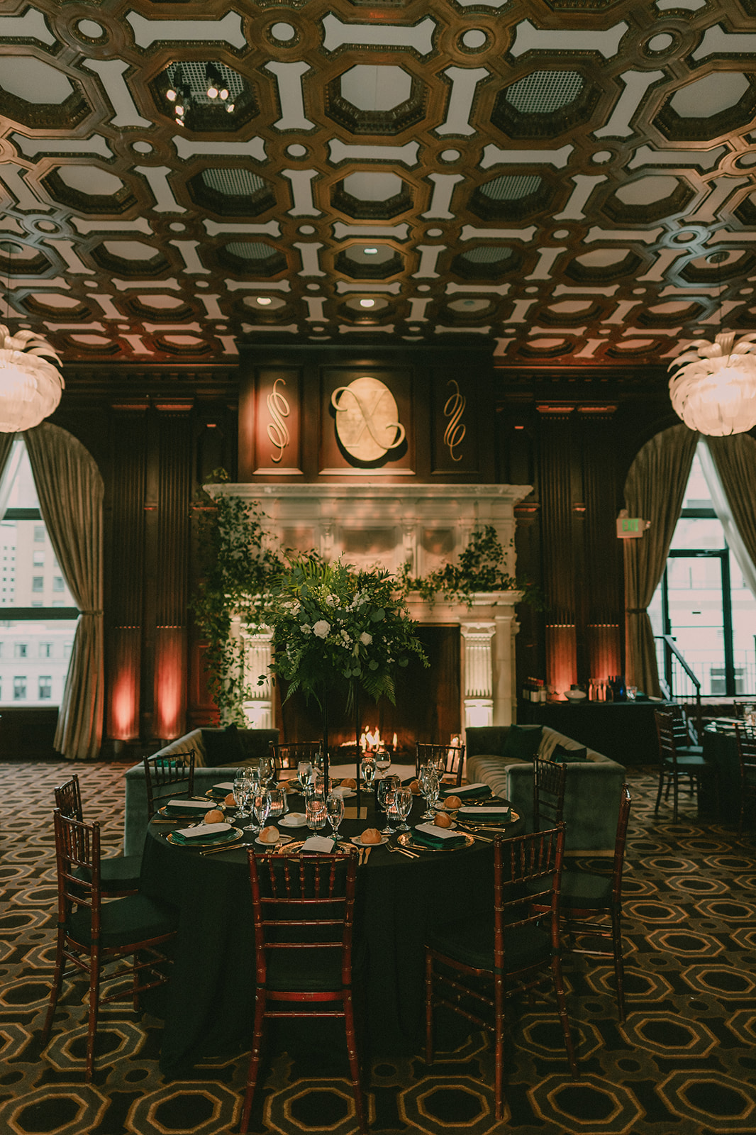 Candlelit reception table beside a marble fireplace beneath an ornate geometric ceiling.