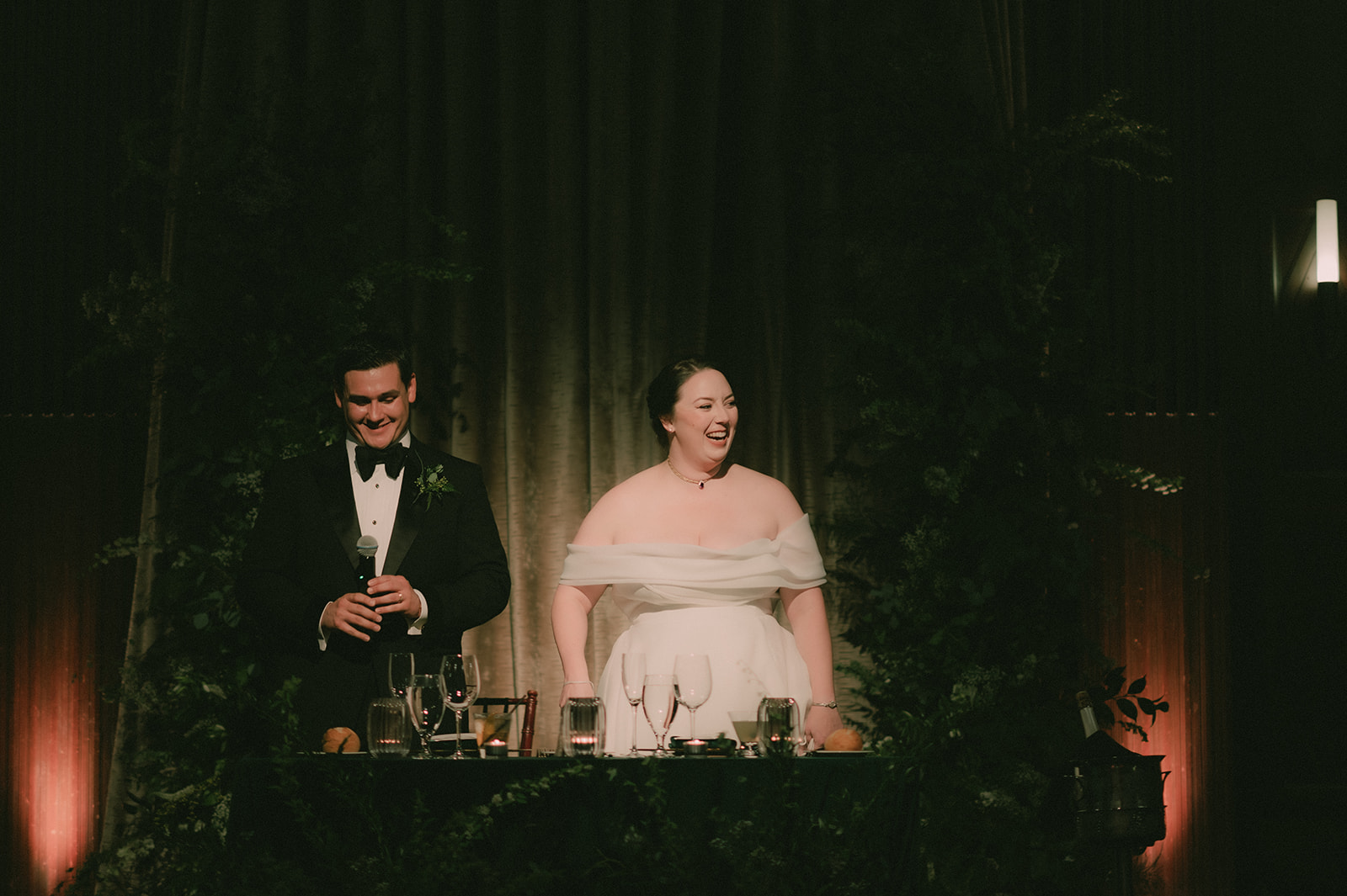 Anne and Charles laugh together at their greenery-covered sweetheart table.