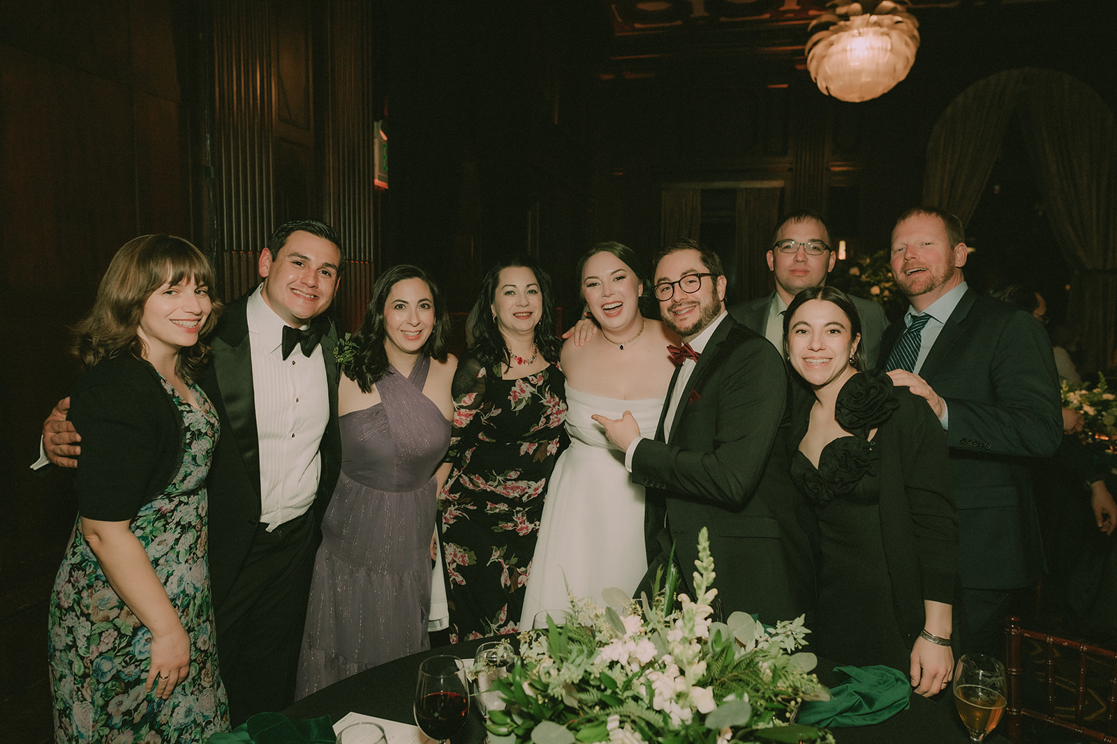 Anne and Charles gather with smiling friends around a flower-filled reception table.
