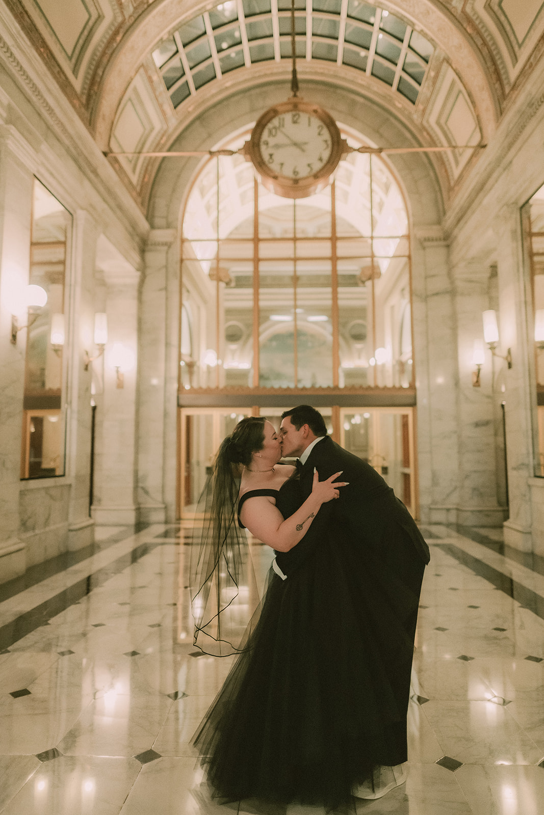 Anne and Charles kiss in the marble lobby beneath the Julia Morgan Ballroom clock.