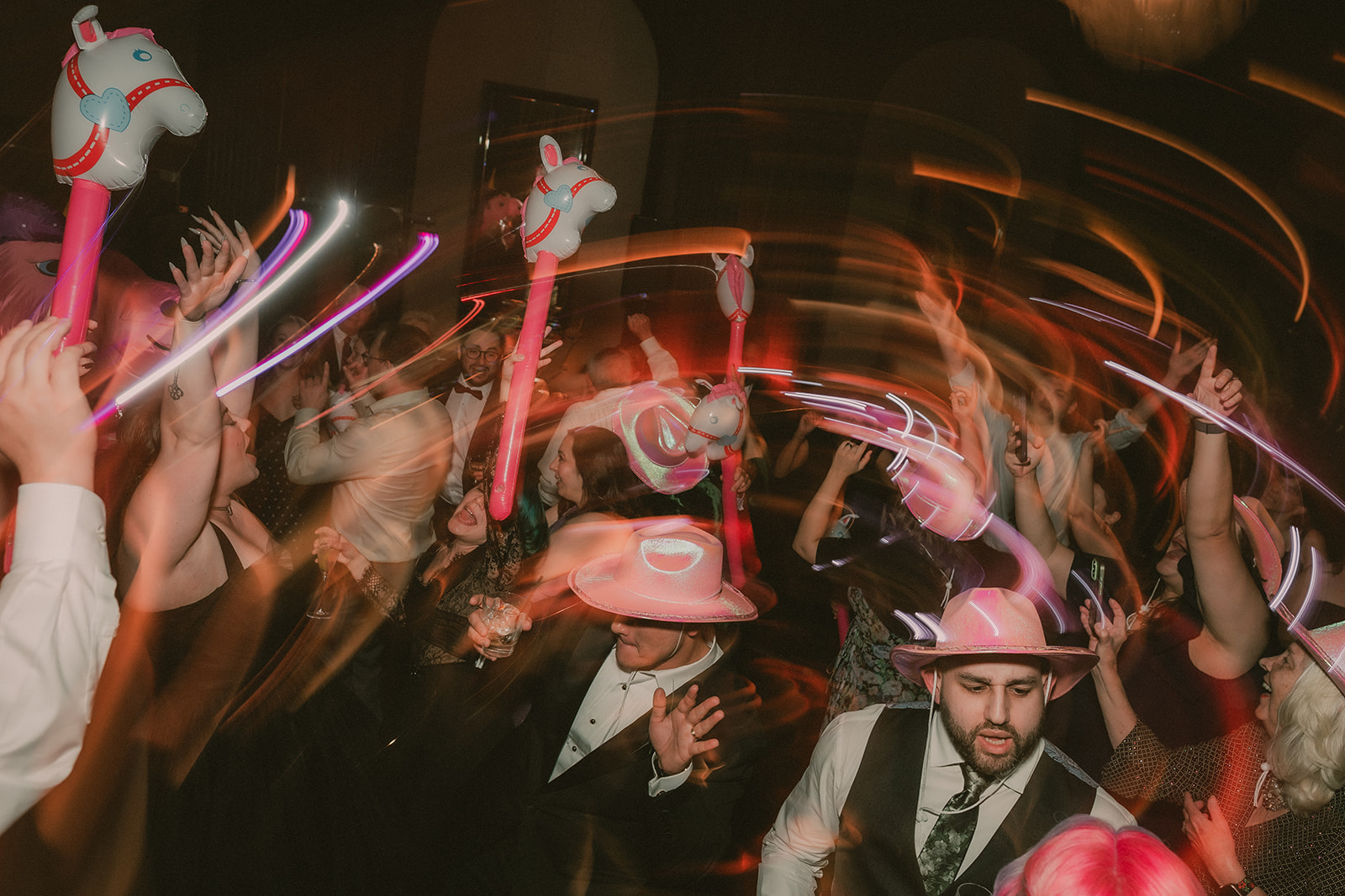 Wedding guests dance with pink pony props beneath sweeping trails of colorful motion blur.