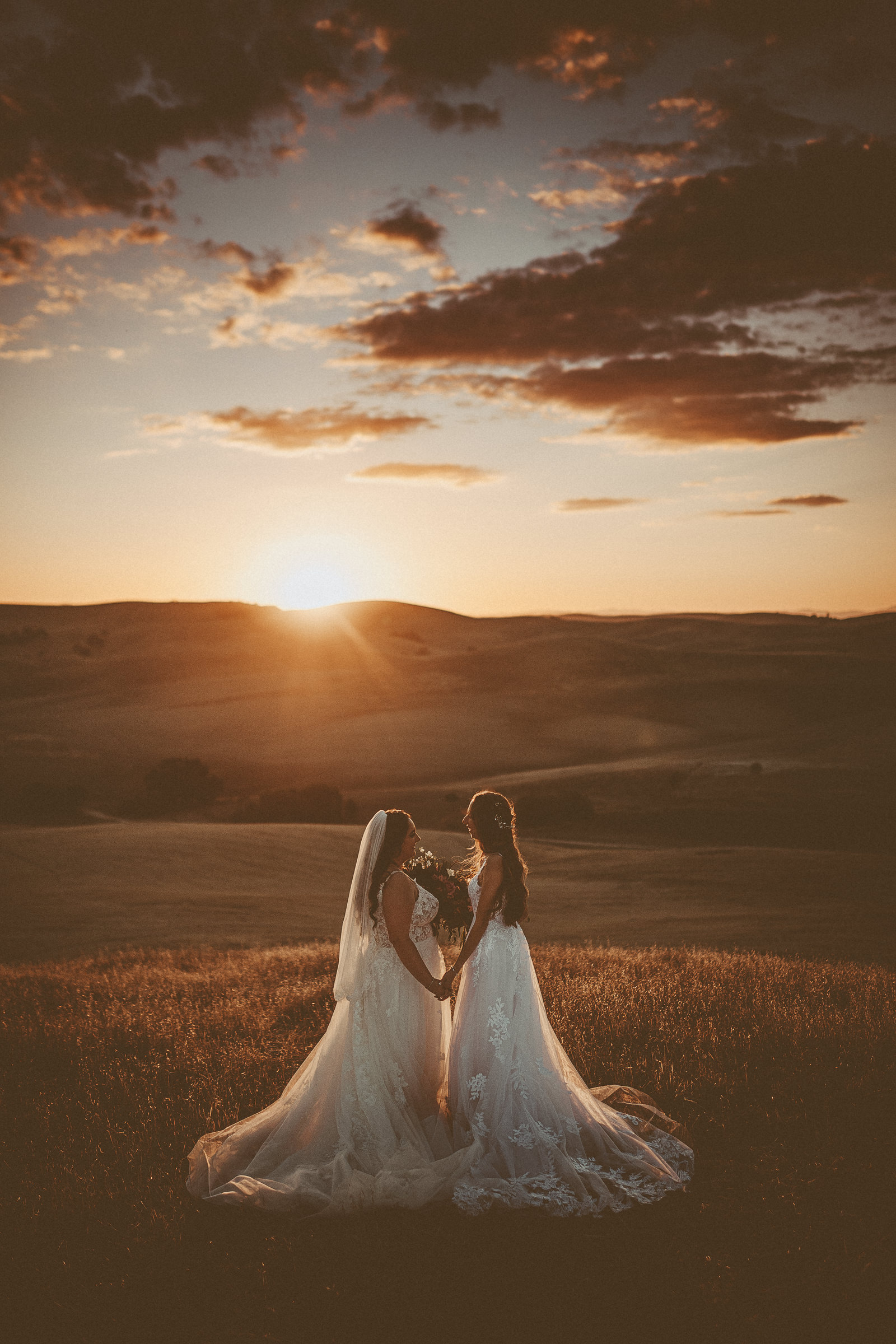 Olivia and Felicity holding hands in flowing wedding gowns beneath a dramatic sunset at Almond Springs