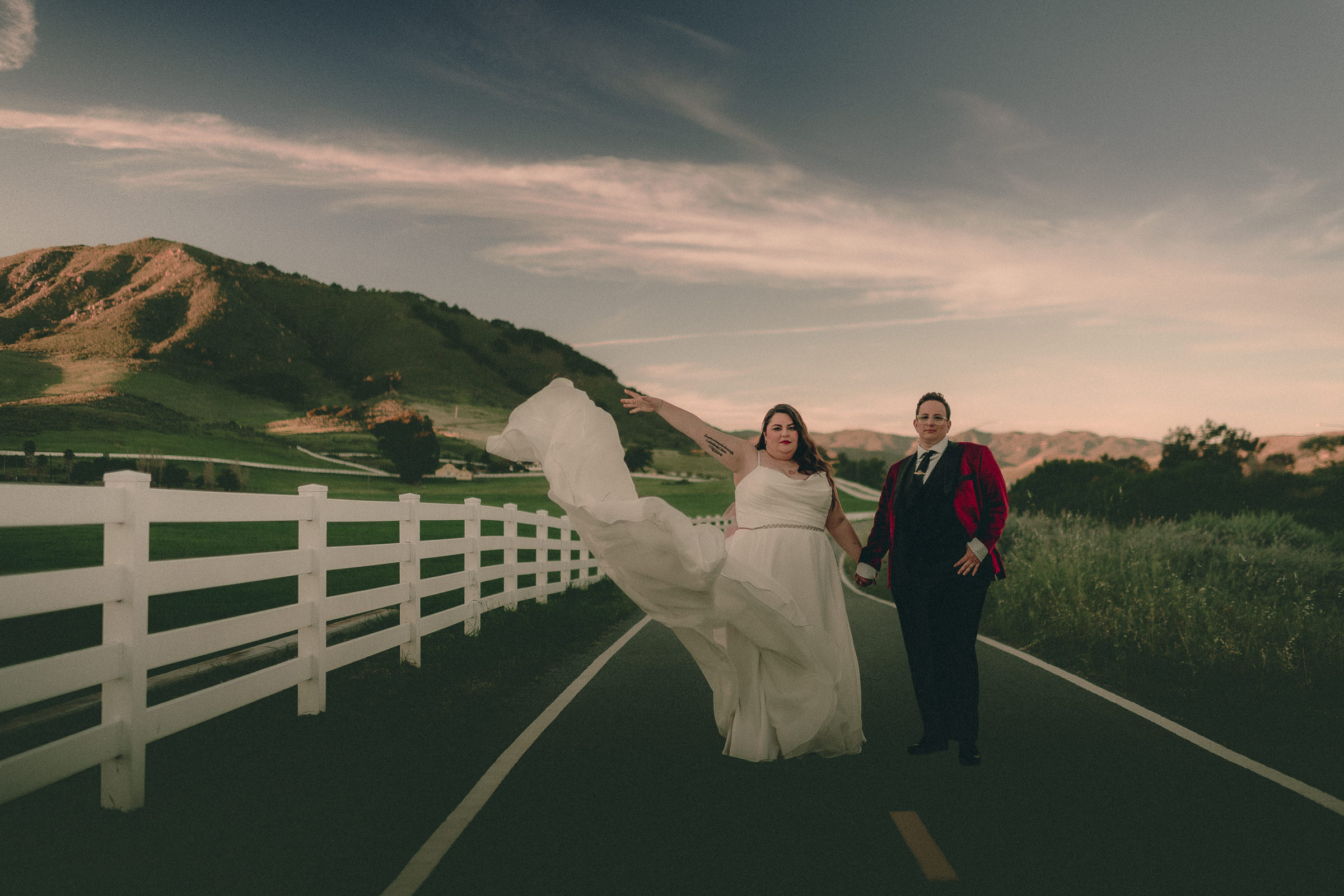 A couple hold hands on a country road at sunset, with a flowing wedding dress, burgundy jacket, white fence and rolling hills.