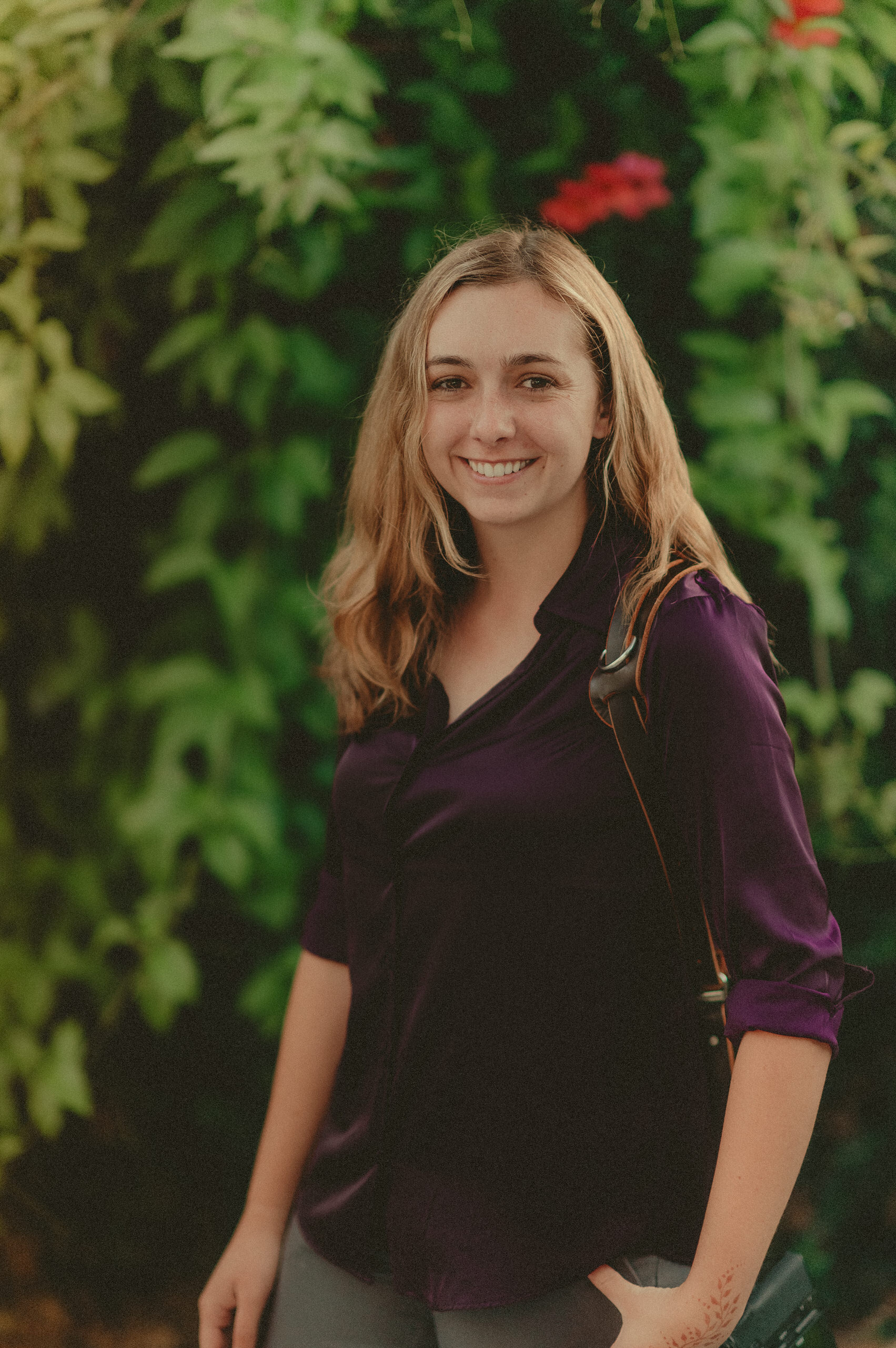 Makayla smiling outdoors in a purple top with camera straps over her shoulders.