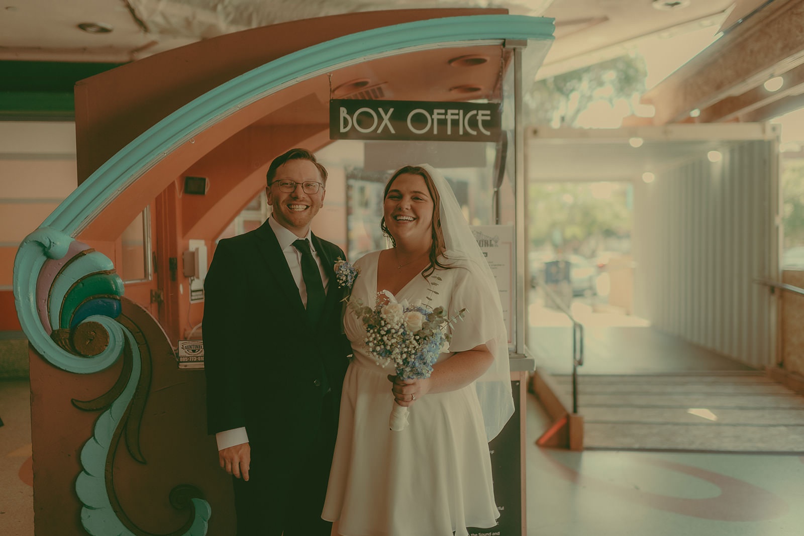 Tiffani and Braden smiling beside a vintage theater ticket booth in downtown San Luis Obispo on their wedding day